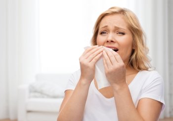 people, healthcare, rhinitis, cold and allergy concept - unhappy woman with paper napkin sneezing over living room background