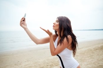 summer, travel, technology and people concept - sexy young woman taking selfie with smartphone and sending blow kiss on beach