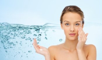 beauty, people, moisturizing cosmetics, skin care and health concept - young woman applying cream to her face over water splash background