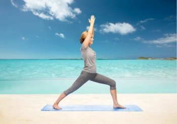 fitness, sport, people and healthy lifestyle concept - woman making yoga warrior pose on mat over beach background