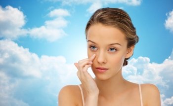 beauty, people, cosmetics, skincare and health concept - young woman applying cream to her face over blue sky and clouds background
