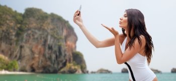 summer, travel, technology and people concept - sexy young woman taking selfie with smartphone and sending blow kiss over rock on bali beach background