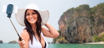 lifestyle, leisure, summer, technology and people concept - smiling young woman or teenage girl in sun hat taking picture with smartphone on selfie stick over rock on bali beach background