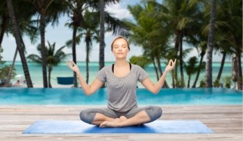 fitness, sport, people and healthy lifestyle concept - woman making yoga meditation in lotus pose on mat over hotel resort pool on tropical beach background