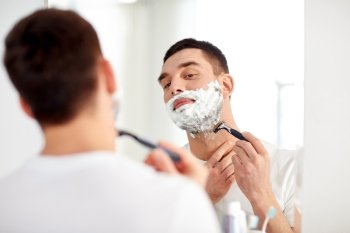 beauty, hygiene, shaving, grooming and people concept - young man looking to mirror and shaving beard with manual razor blade at home bathroom