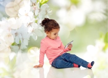 childhood, technology and people concept - smiling little african american baby girl playing with smartphone and sitting on floor over cherry blossoms background