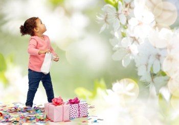 childhood, birthday, party, holidays and people concept - happy little african american baby girl with gift boxes and confetti playing with shopping bag looking up over cherry blossoms background