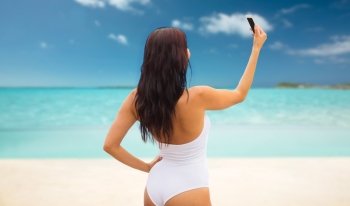 summer, travel, technology and people concept - sexy young woman taking selfie with smartphone over tropical beach background