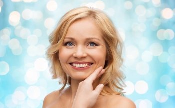 beauty, people and skincare concept - smiling woman with bare shoulders touching face over blue holidays lights background
