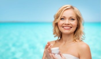 beauty, people, skincare and cosmetics concept - happy middle aged woman with cream jar over blue sea and sky background