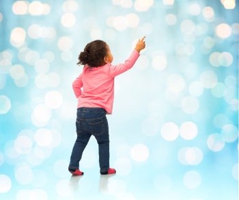 people, race, ethnicity and childhood concept - happy african american baby girl pointing finger to something from back over blue holidays lights background