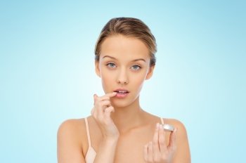 beauty, people and lip care concept - young woman applying lip balm to her lips over blue background