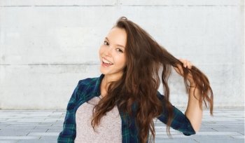 people, hair care, style and teens concept - happy smiling pretty teenage girl strand of hair over gray urban street background