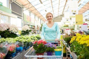 people, gardening, sale and consumerism concept -happy woman with shopping cart and flowers in shop or greenhouse