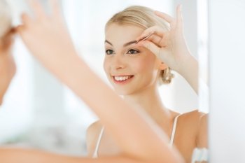 beauty and people concept - smiling young woman with tweezers tweezing eyebrow and looking to mirror at home bathroom. woman with tweezers tweezing eyebrow at bathroom