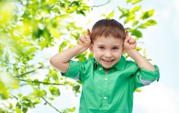 childhood, fashion, fun, summer and people concept - happy little boy having fun and making horns over green natural background