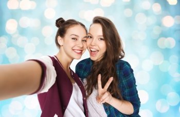 people, friends, teens and friendship concept - happy smiling pretty teenage girls taking selfie and showing peace sign over blue holidays lights background