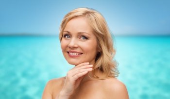 beauty, people and skincare concept - smiling middle aged woman with bare shoulders touching face over blue sea and sky background