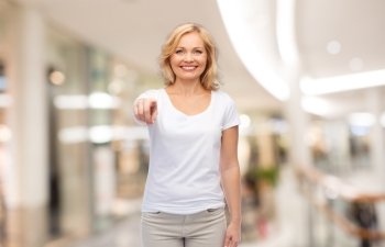 gesture, advertisement and people concept - smiling woman in blank white t-shirt pointing finger to you at shopping center or mall