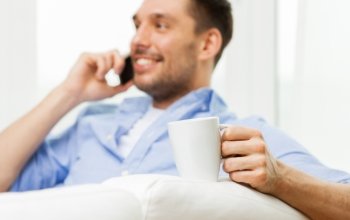 drinks and people concept - close up of smiling man with cup of coffee or tea calling on smartphone at home. smiling man with cup calling on smartphone at home