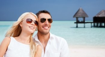 summer holidays, tourism, vacation, travel and dating concept - happy couple in shades at sea side over beach with bungalow background