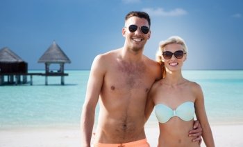 love, travel, tourism, summer and people concept - smiling couple on vacation in swimwear and sunglasses hugging over beach with bungalow background