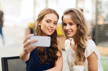 technology, lifestyle, friendship and people concept - happy young women or teenage girls with smartphone and coffee cups taking selfie at outdoor cafe