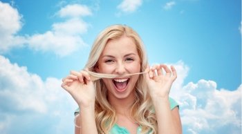 emotions, expressions, hairstyle and people concept - smiling young woman or teenage girl making mustache with strand of hair over blue sky and clouds background