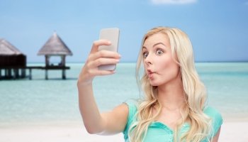 expressions, technology, travel, tourism and people concept - funny young woman or teenage girl taking selfie with smartphone and making fish face over tropical beach with bungalow background
