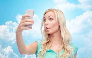 expressions, technology and people concept - funny young woman or teenage girl taking selfie with smartphone and making fish face over blue sky and clouds background