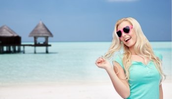 travel, tourism, summer vacation and people concept - smiling young woman or teenage girl in sunglasses holding her strand of hair over tropical beach with bungalow background