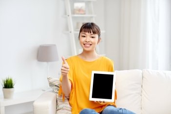 people, technology, gesture, advertisement and leisure concept - happy young asian woman sitting on sofa and showing tablet pc computer blank black screen and thumbs up at home
