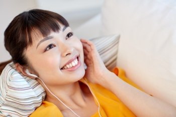 people, technology and leisure concept - happy asian young woman lying on sofa with earphones listening to music at home