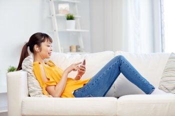 people, technology and leisure concept - happy asian young woman sitting on sofa with smartphone and earphones listening to music at home