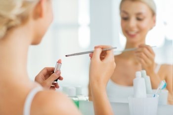 beauty, make up, cosmetics, morning and people concept - close up of smiling young woman with lipstick and make up brush at home bathroom mirror