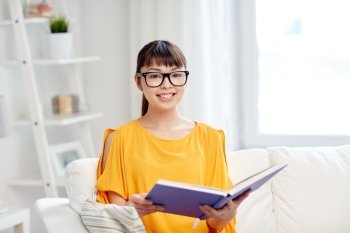 education, knowledge, leisure, literature and people concept - smiling young asian woman or student girl in glasses reading book at home