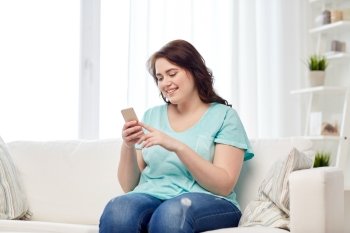 people, technology, communication and leisure concept - happy young plus size woman sitting on sofa with smartphone at home