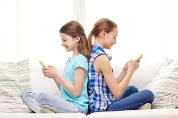people, children, technology, friends and friendship concept - happy little girls with smartphones sitting on sofa back to back at home
