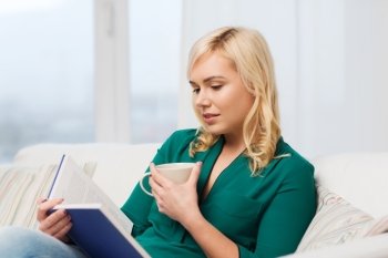 leisure, literature and people concept - young woman with tea cup reading book at home