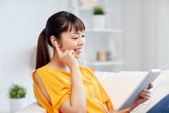 people, technology and leisure concept - happy asian young woman sitting on sofa with tablet pc computer and earphones listening to music at home