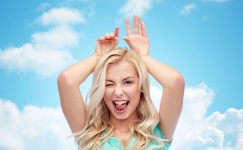 fun, expressions, easter and people concept - happy smiling young woman making bunny ears over blue sky and clouds background
