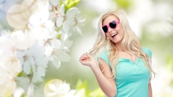 emotions, expressions, summer and people concept - smiling young woman or teenage girl in sunglasses holding her strand of hair over natural spring cherry blossom background