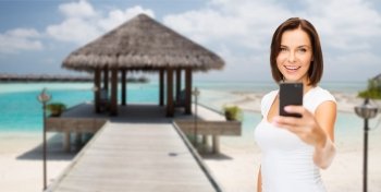 travel, tourism, summer vacation, technology and people - happy young woman taking selfie picture by smartphone on maldives beach with bungalow background