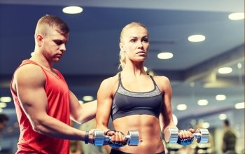 fitness, sport, bodybuilding and weightlifting concept - young woman and personal trainer with dumbbells flexing muscles in gym