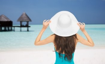 people, summer holidays, travel, tourism and vacation concept - woman in swimsuit and sun hat from back over maldives beach with bungalow background