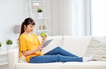 people, healthy eating, education, technology and concept - happy young asian woman sitting on sofa with tablet pc computer and green apple at home