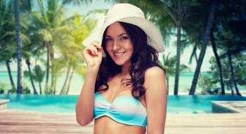 people, fashion, travel, tourism and summer concept - happy young woman in bikini swimsuit and sun hat over swimming pool and beach with palm trees background