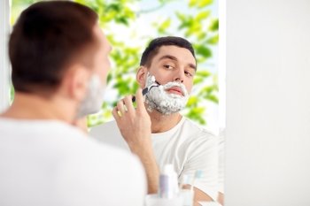 beauty, hygiene, shaving, grooming and people concept - young man looking to mirror and shaving beard with manual razor blade at home bathroom over green natural background