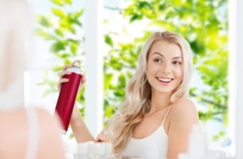 beauty, hygiene, hairstyle, morning and people concept - smiling young woman with hairspray styling her hair and looking to mirror at home bathroom over green natural background