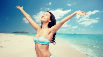 people, tourism, travel and summer concept - happy young woman in bikini swimsuit with raised hands over beach background